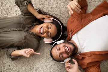 Cheerful Black Couple Wearing Headphones Listening To Music Lying Indoors