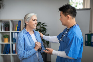 Happy old caucasian female patient at meeting thanks to millennial japanese doctor in uniform in office