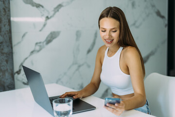 Confidentwoman remote employee freelancer sit at kitchen table search files on phone to forward on laptop via home wireless network. Smart young lady user sync restore data using two gadgets