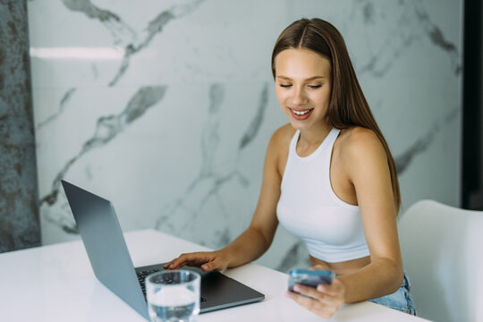 Confidentwoman Remote Employee Freelancer Sit At Kitchen Table Search Files On Phone To Forward On Laptop Via Home Wireless Network. Smart Young Lady User Sync Restore Data Using Two Gadgets