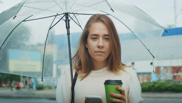 joyful girl drinks tea on the street under an umbrella in rainy weather, and speaks on the phone.