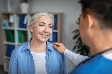 Obraz premium Cheerful senior european lady patient on meeting with young chinese doctor in uniform talking in office