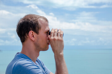A young man looks through binoculars at the blue sea. Travel, rest, adventure.