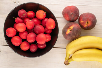 A few sweet juicy cherry plums, three peaches and a bunch of bananas with a black ceramic plate on a wooden table, close-up, top view.