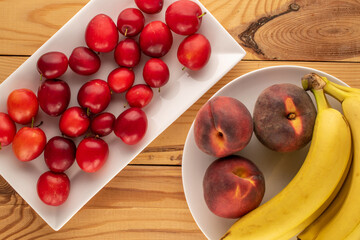 Several sweet juicy cherry plums, three peaches and a bunch of bananas with white ceramic utensils on a wooden table, close-up, top view.