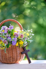 Christians cross and meadow flowers in wicker basket on table, blurred green natural background....