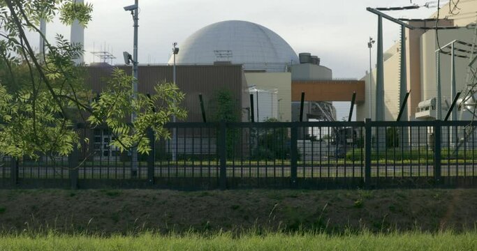 Nuclear reactor building of a nuclear power station behind a fence with grass of a meadow and trees in the foreground. Transformer on the right side.