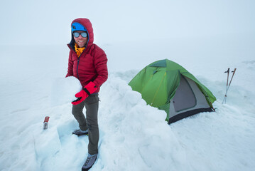 Smiling man dressed red warm mountaineering down jacket making the protective wall from snow bricks...