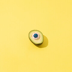 Sliced avocado with plastic eye instead of stone on yellow background. Happy halloween concept with copy space.