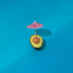 Slice of avocado with avocado stone and pink cocktail umbrella on blue background with copy space. Summer vacation concept.