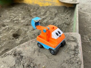 Closeup of the orange toy excavator with a blue bucket in a sandbox. A small children's excavator with kind eyes and smile left in the sand in a sandbox