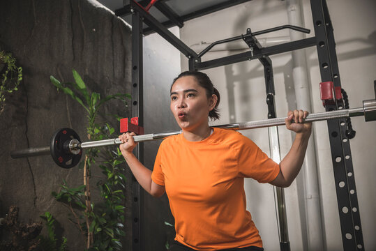 A determined asian woman exhaling properly via the mouth while performing a set of barbell squats from a home workstation. Pursed lip breathing.