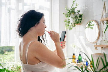 Young beautiful woman with overweight brushing her teeth and looking at the phone in light batroom with many green plants. Wellness, everyday technology