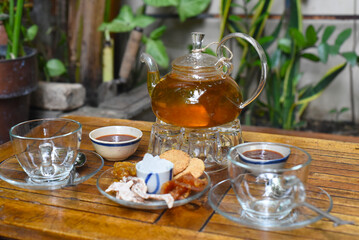 Glass tea pot with cups of marmalade and crystalized fruits on wooden table 