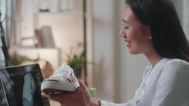 Close Up Of Asian Female Footwear Designer Looking At The Colourful Pattern Sneaker In Hands And Comparing It To The Pictures On A Laptop At Home. Shoe Production Procedure Concept 
