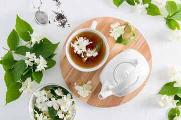 top view of a cup with fragrant jasmine tea and a white ceramic teapot on a round wooden board. white background with flowers and jasmine leaves.