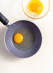 Raw eggs in a frying pan and raw egg in glass bowl. Top view. Broken white background.