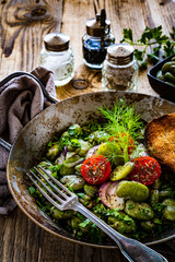 Broad beans with cherry tomatoes, garlic, toasted bread and parsley in cooking pan on wooden table
