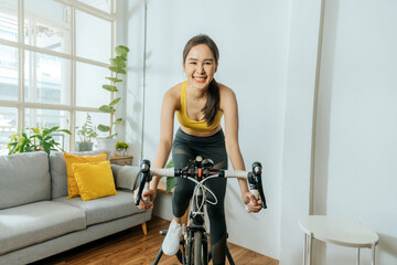 Fototapeta na wymiar athletic asian young slim body woman wearing sportswear looking at camera and exercise cyclist on bicycle in living room at home for good healthy, bodybuilder, lifestyle, sport, home exercise concept