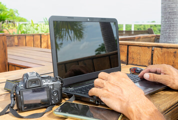 A photographer works with a laptop computer on the wooden garden table on a sunny day. Selective focus