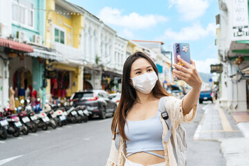 Asian traveler wearing a medical mask take a photo or video call with her family and smiles while...