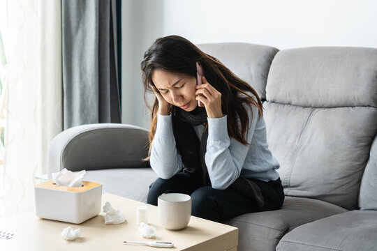 Sick Asian woman sitting on sofa in living room at home and talking with doctor or her family on mobile phone for counseling. Cold, flu, home isolation during coronavirus pandemic  concept. Closeup