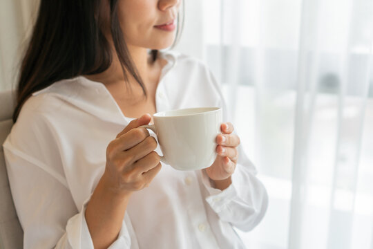 Young beautiful brunette hair woman in white shirt drinking coffee while sitting on the chair and look out the  window in the morning. Copy space, closeup