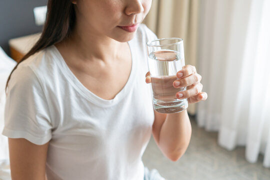 Healthy young Asian woman smiling and enjoying fresh clean water while sitting on bed in morning. Diet girl drinking water after wake up. Concept of health and freshness, thirst, water purification