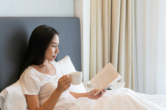 Young beautiful Asian woman in white T-shirt pajamas drinking coffee while reading a book sitting on the bed in the morning. Copy space