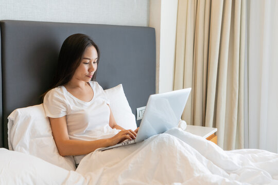 Happy beautiful Asian businesswoman working on a laptop while sitting on bed at home. Work from home. Technology and lifestyle concept. Closeup, copy space.
