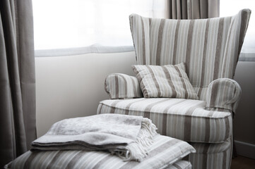 Gray wing chair, puff and blanket placed next to a window inside a house. Furniture for decorating. Color combination.