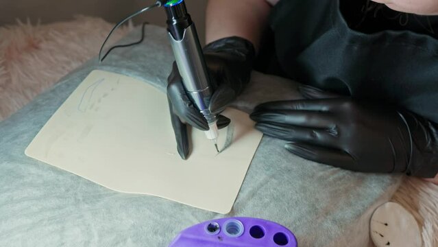 a young woman in black protective gloves with a permanent makeup machine makes a test eyebrow dyeing on a silicone mat, picks up paint and then works with a machine. permanent makeup master training