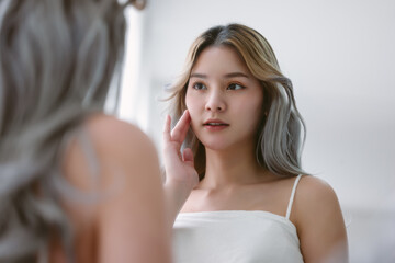 Beautiful young asian woman looking at mirror touching her skin enjoying treatment for skin. Korean...
