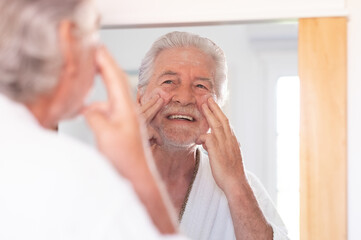 Skin care. Handsome senior man applying cream at his face and looking at himself with smile while...