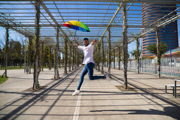 Handsome young blue-eyed gay man is jumping with open arms and holding a rainbow-coloured umbrella...
