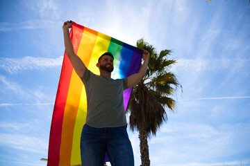 Handsome young gay man holding the gay pride flag in the wind, blue sky in the background. The flag...