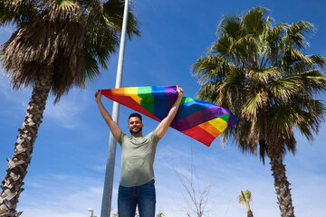 Handsome young gay man holding the gay pride flag in the wind, blue sky in the background. The flag flutters in the wind. Concept homosexuality, equality, rights. 28 June