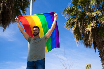 Handsome young gay man holding the gay pride flag in the wind, blue sky in the background. The flag...