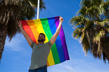 Handsome young gay man holding the gay pride flag in the wind, blue sky in the background. The flag...