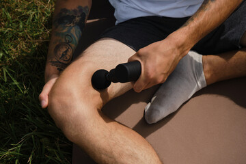 Concept of healthy lifestyle. Athlete stretches leg muscles after training using electronic body machine sitting on sports mat in park. Caucasian man massages himself with percussion gun outside.