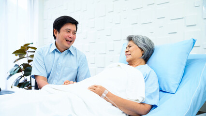 Obraz premium Son of an elderly woman patient come to see his mother treated at the hospital. Female patient lying in bed receiving saline in the patient room. Smile brightly when encouraged.
