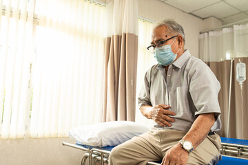 Asian senior male patient sitting on examination bed pointing at the stomach that pain to a...