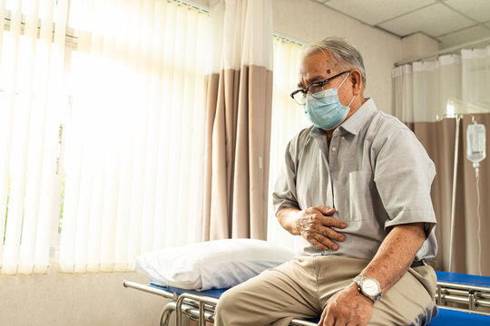 Asian Senior Male Patient Sitting On Examination Bed Pointing At The Stomach That Pain To A Caucasian Male Doctor.Elder Man Showing Stomach Ache Pain To Doctor In Clinic.