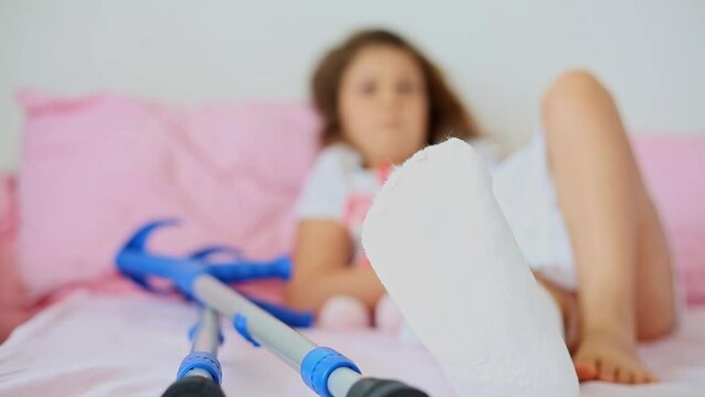 Unrecognizable little girl with a broken leg at home on a bed with crutches lying nearby. Fractured leg in a cast in a young child. Childhood trauma