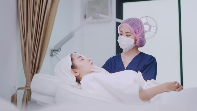 Asian female beautician in blue clothes, medical hat, and face mask doing facial peeling procedure with microdermabrasion machine to Asian female client lying on bed in treatment room of beauty clinic