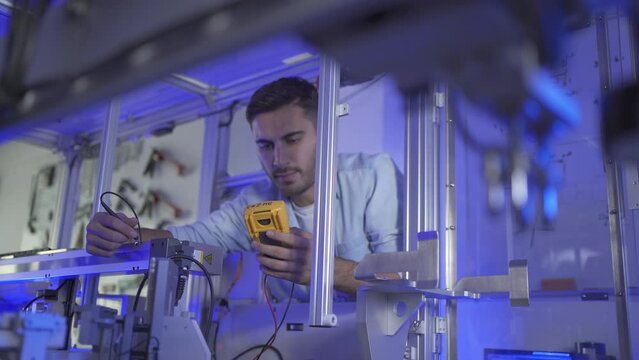 Young Caucasian male electrical engineer measuring the voltage on the machine and looking at a display on a digital multimeter, to adjust an electrical system for the machine in an industrial workshop