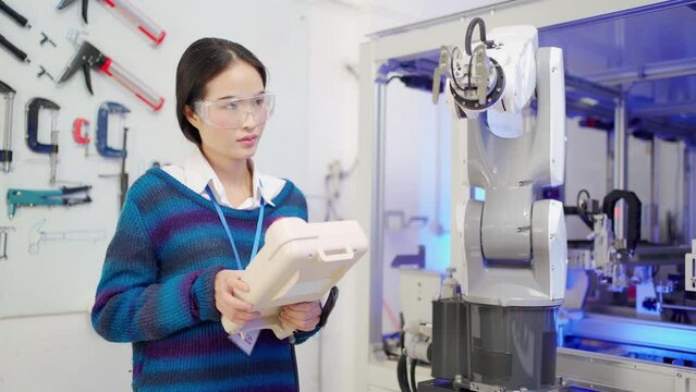 A beautiful Asian female apprentice engineer wearing goggles standing using a robot controller, testing and controlling the operation of an automation robotic arm machine in an industrial workshop.