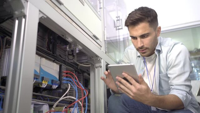 Young Caucasian male electrical engineer in a blue shirt squatting checking wire cables that is plugged on the back of the machine with the digital tablet in his hand in an industrial factory.