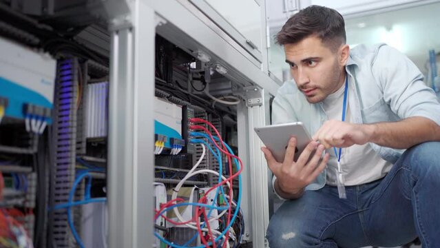 Young Caucasian male electrical engineer in blue shirt squatting behind the machine using a digital tablet checking wire cables and the electrical system of the machine in an industrial factory.