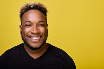 Obraz premium A young stylish black man in an everyday black T-shirt smiles against a yellow background at a close-up camera. Curly African-American shines with a snow-white smile after the dentist
